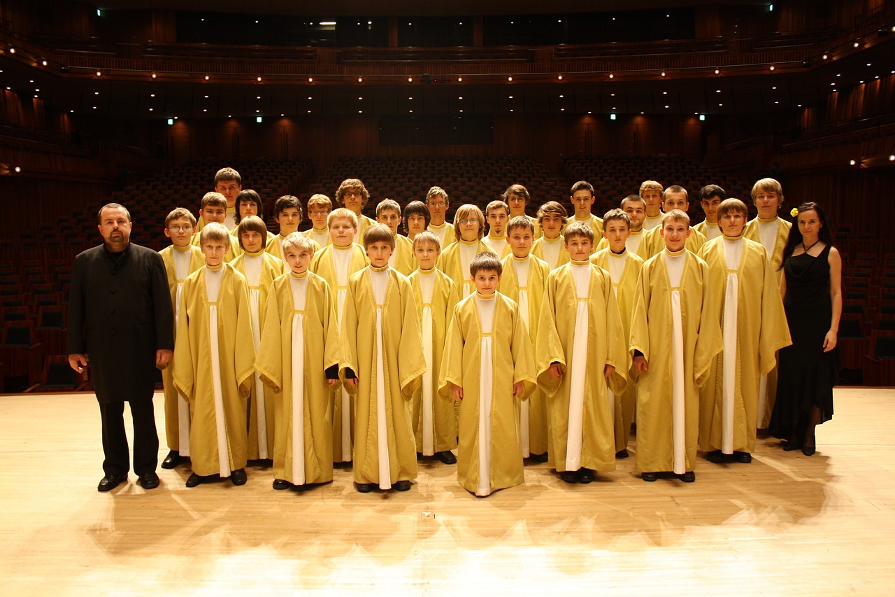 A choir group on stage in a theater setting, wearing gold robes, under dim lighting.